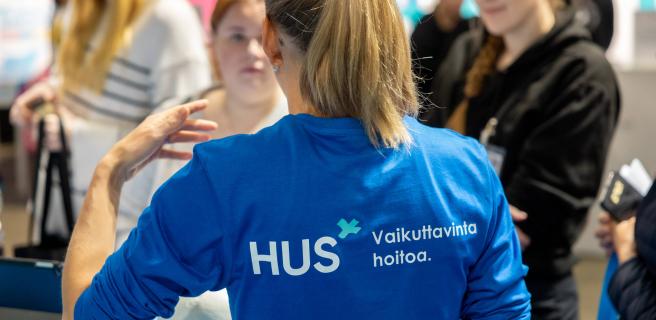 HUSin työntekijä messustandilla esittelemässä. Selin. Paidassa lukee Vaikuttavinta hoitoa.