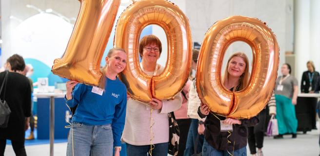 Kolme naista pitelee folioilmapalloja, joista muodostuu luku sata.