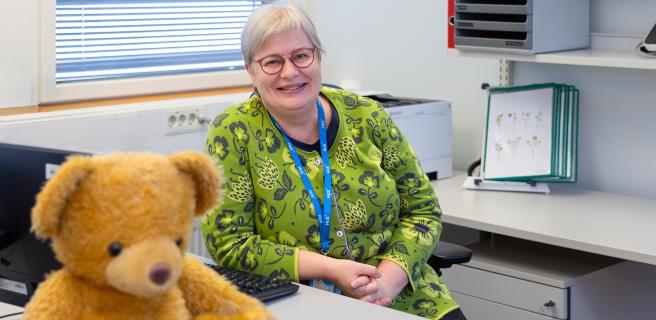 HUSin työterveyshoitaja Tarja Siik istuu työpöydän ääressä pehmonalle kuvan etualalla.