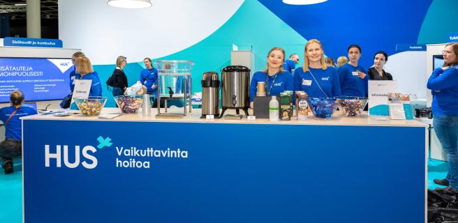 Messutiskillä kahvia ja suklaata. Messutiskin takana kaksi HUSin messuedustajaa ja yleisöä