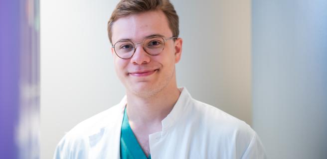 A person wearing glasses and a white lab coat, smiling in a well-lit room.