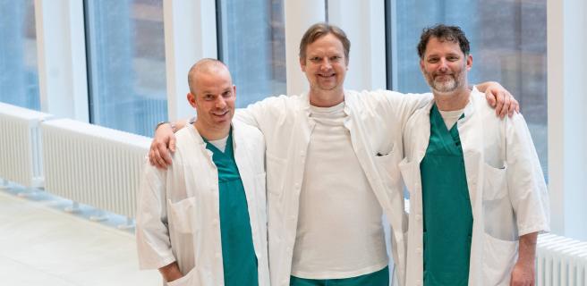 Kolme lääkäriä seisoo yhdessä hymyillen käytävällä sairaala-asuihin pukeutuneina.