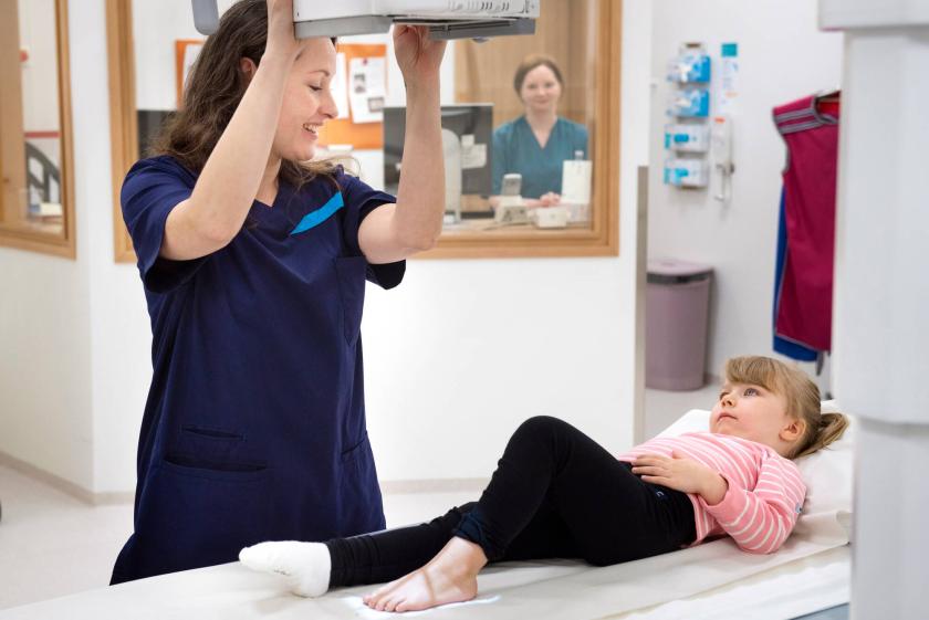 Lapsi makaa tutkimuspöydällä ja hoitaja valmistelee jalan röntgenkuvausta.