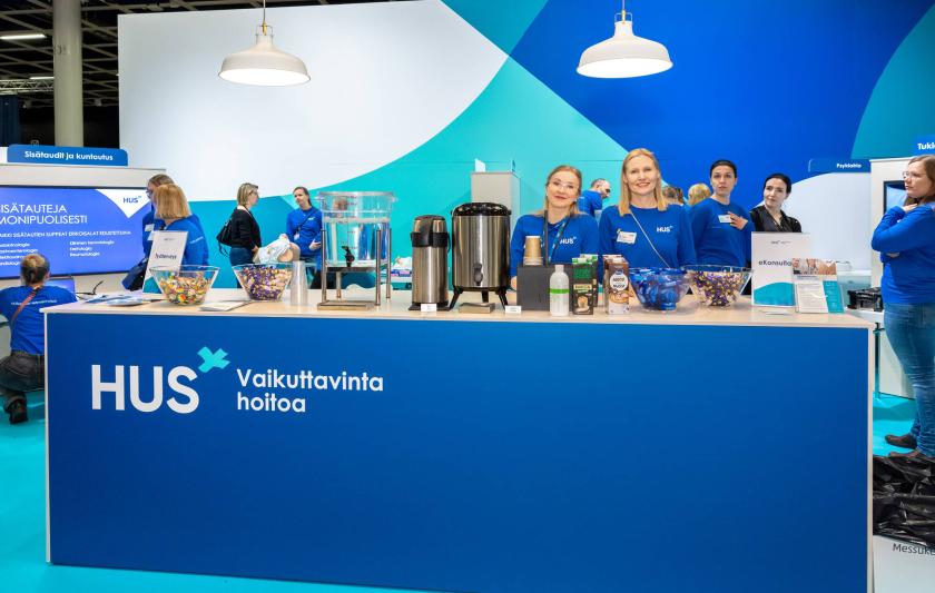 Messutiskillä kahvia ja suklaata. Messutiskin takana kaksi HUSin messuedustajaa ja yleisöä