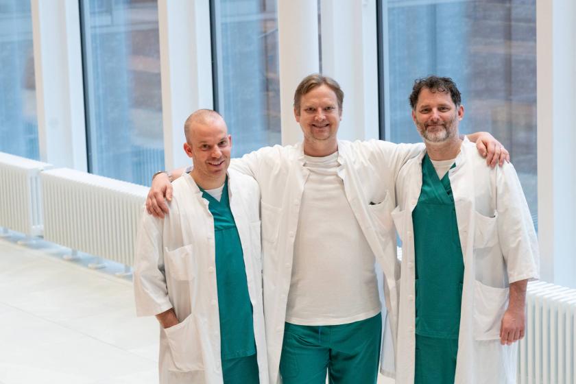 Kolme lääkäriä seisoo yhdessä hymyillen käytävällä sairaala-asuihin pukeutuneina.