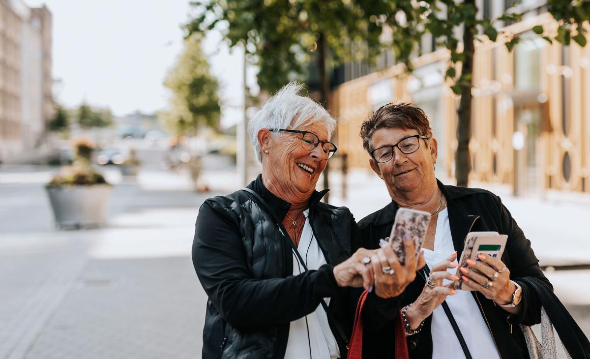 Två äldre personer som antas vara kvinnor står på gatan med mobiltelefoner i händerna och ser på den ena personen som antas vara kvinnas mobiltelefon.
