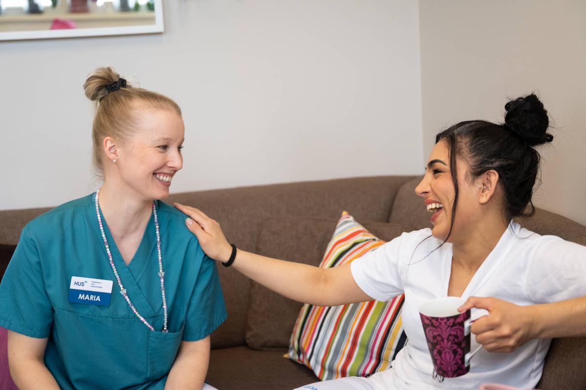 Kaksi hymyilevää hoitajaa, joista toinen koskettaa toista olkapäähän.