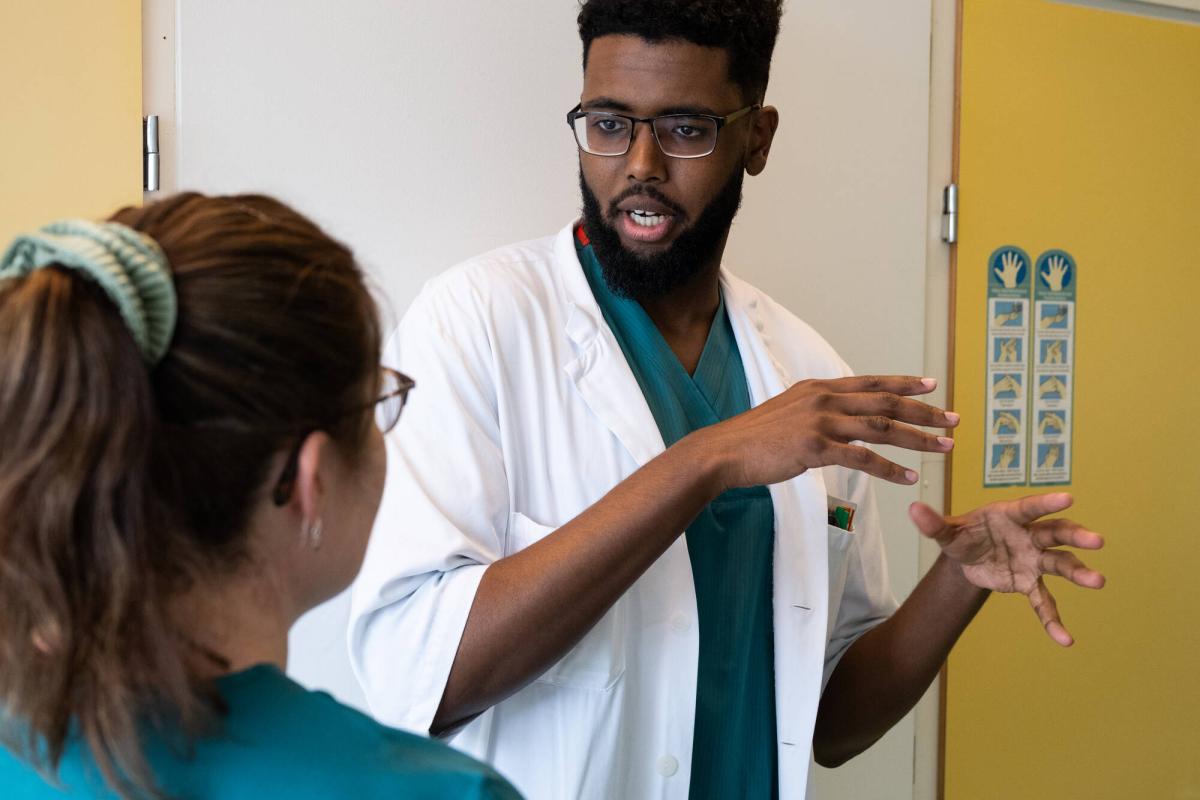 Two healthcare professionals are discussing in a corridor. One of them is wearing a white coat and illustrating something with hand gestures.