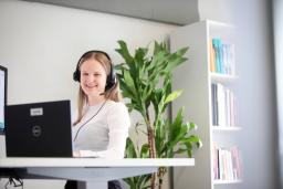 Hymyilevä asiantuntija työskentelee tietokoneella kuulokemikrofoni päässä valoisassa toimistoympäristössä.