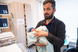 Mies pitelee sylissään vastasyntynyttä vauvaa sairaalahuoneessa. Vauva on kääritty vaaleansiniseen peittoon ja hänellä on sinivihreä pipo. Taustalla näkyy sairaalalaitteita ja ikkuna.