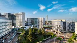Bild av Mejlans campus med flera byggnader, omgivna av träd och parkeringsplatser under en klarblå himmel.