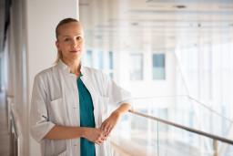 Henkilö valkoisessa takissa seisoo käytävällä ja hymyilee.